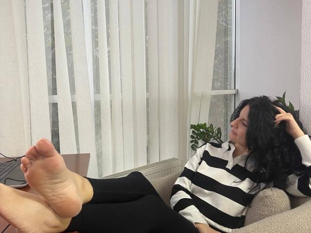 Marlinaa with black hair, white and black striped shirt, and black pants, sitting with feet up, looking to the side, on a beige couch, hand in hair.