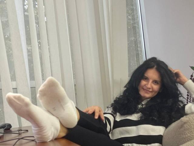 Marlinaa with long black curly hair, smiling, wearing a black and white striped sweater, black pants, and white socks, lying on a couch with her feet up.