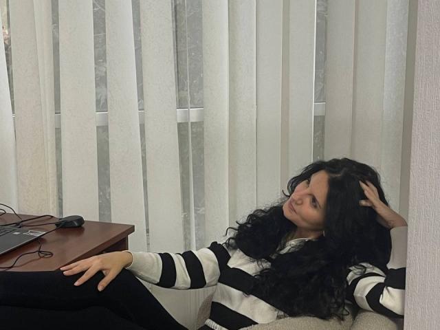 Marlinaa with long black curly hair, wearing a black and white striped shirt, sitting on a chair, hand in hair, looking bored, black pants, laptop and cord on wooden table.
