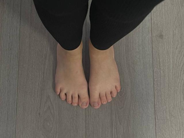 Marlinaa's bare feet standing on wooden floor, black pants covering legs, toes slightly curled, skin tone light, small toenails, wood texture visible, perspective from above.