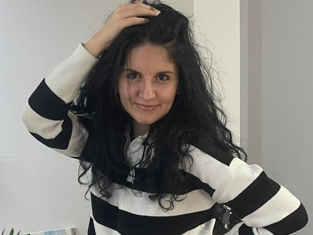 Marlinaa with black curly hair, smiling, wearing a black and white striped sweater, hand in her hair, sitting, white wall in background. Boobs not visible.