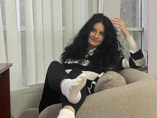Marlinaa with long black curly hair, smiling, wearing a black and white striped sweater and black pants, sitting on a beige couch, legs up, white socks, right hand raised.