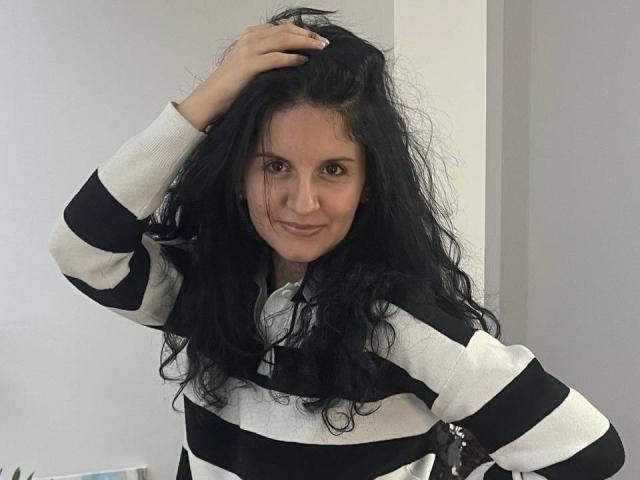 Marlinaa with curly black hair, wearing a black and white striped sweater, hand in hair, smiling, brown eyes, light skin, standing in a room, looking at camera.