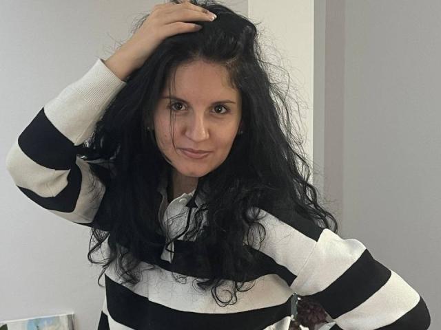 Marlinaa with messy black hair, wearing a white and black striped sweater, hand in hair, smiling, brown eyes, light skin, medium build, slight smirk, casual indoor setting.