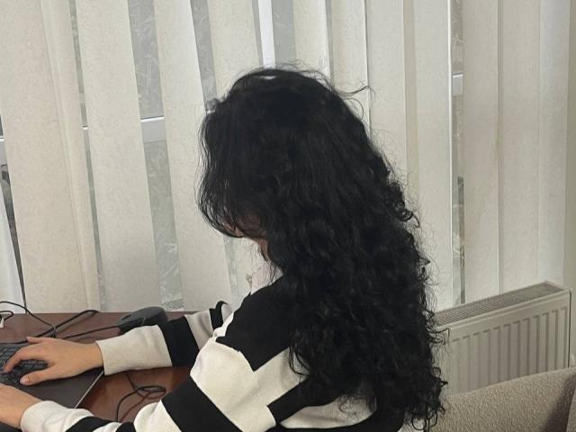 Marlinaa with long, curly black hair, wearing a black and white striped shirt, sitting at a desk, using a keyboard with a mouse in front of her.