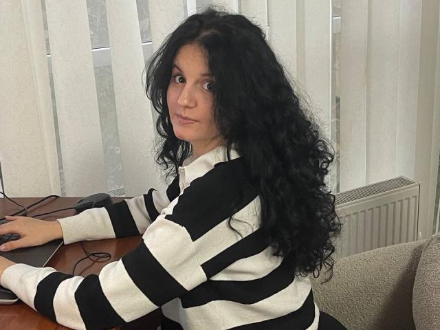 Marlinaa with long curly black hair, wearing a black and white striped sweater, sits at a wooden table, looking over her shoulder with a slight smile, hands on a mouse.