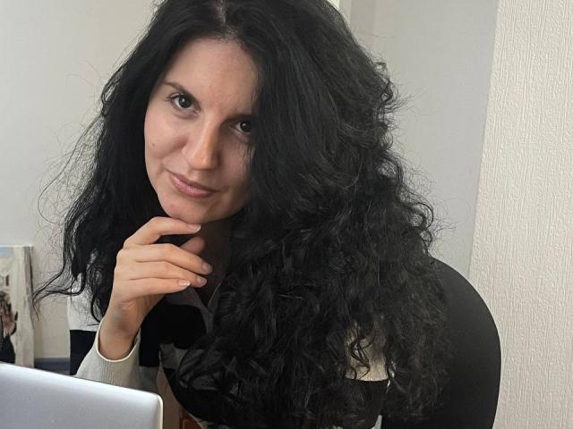 Marlinaa with long, curly black hair, light skin, and brown eyes, slightly smiling, touching her chin with her right hand, wearing a black and white top, in front of a laptop.