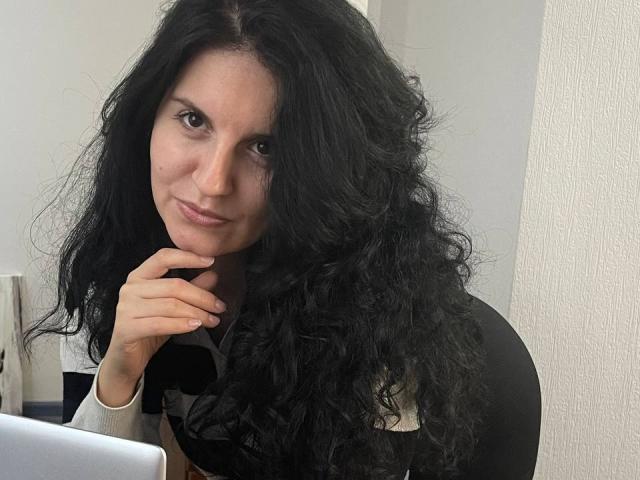 Marlinaa with curly black hair, brown eyes, and a subtle smile, resting her chin on her hand, wearing a black top, in front of a laptop.