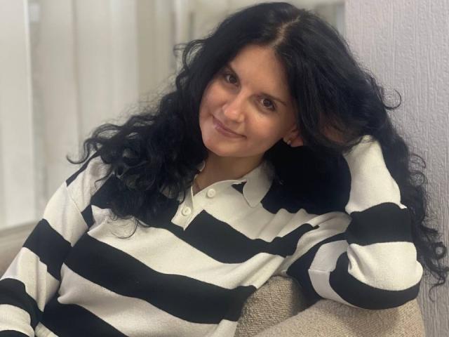 Marlinaa with long black curly hair, smiling, wearing a black and white striped polo shirt, leaning against a wall, hand in her hair.
