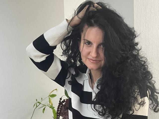 Marlinaa with long, curly black hair, smiling slightly, wearing a black and white striped sweater, hand in hair, standing near a green plant.