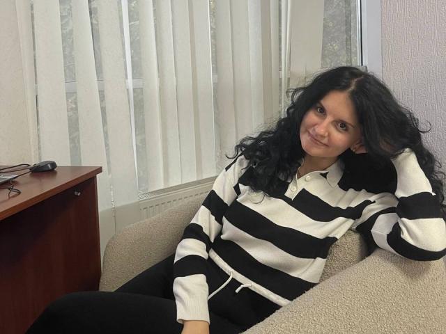 Marlinaa with long black curly hair, wearing a black and white striped hoodie, and black pants, sitting on a beige couch, smiling, hand in her hair, next to a wooden table.
