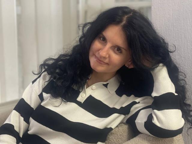 Marlinaa with wavy black hair, wearing a black and white striped polo shirt, leaning against a wall, smiling slightly, looking relaxed and cute.