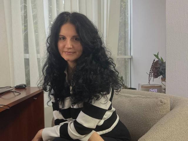 Marlinaa with long curly black hair, smiling, wearing a black and white striped shirt, sitting on a beige couch, with a wooden table and window in the background.