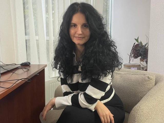 Marlinaa with long, curly black hair, smiling, wearing a black and white striped sweater and black pants, sitting on a beige chair, hands on her knees.