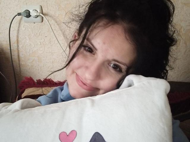 Marlinaa with dark, messy hair, smiling, holding a white pillow with a pink heart, wearing a blue shirt, wall plug in background, slight acne on her cheek.