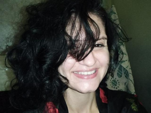Marlinaa with messy black curly hair, smiling with white teeth, wearing a black shirt with red floral patterns, showing her cute face and slightly flushed skin.