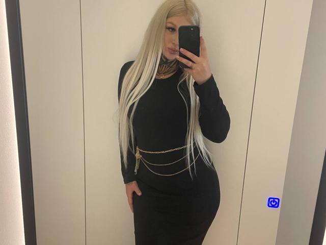 KellyBabbe with long blonde hair, black dress, gold chain belt, and pouty lips, taking a mirror selfie, showing her curvy, round ass and small tits.