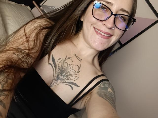 VelvetGreeny with long brown hair, glasses, nose ring, smiling, black top, showing large flower tattoo on chest, and arm tattoos, in a selfie. Boobs visible.