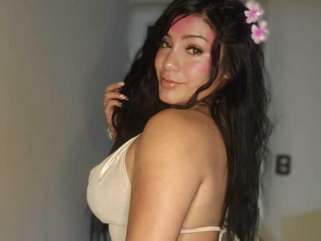 NahomyVolkov with pink makeup on her forehead, black wavy hair, wearing a white tank top, showing her boobs and ass, smiling, holding her hair, pink flowers in her hair.