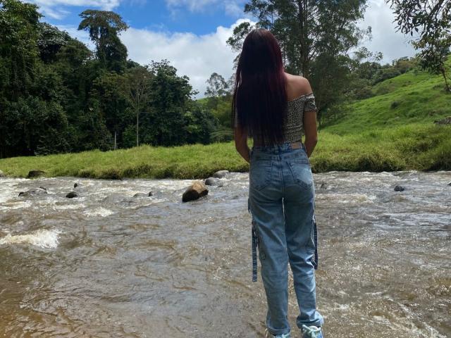 Candy Mendez, com cabelos ruivos longos, blusa com um ombro descoberto e calças jeans de cintura alta, está em um riacho rochoso, de costas, exibindo suas nádegas e calças jeans azuis amarradas nos joelhos..