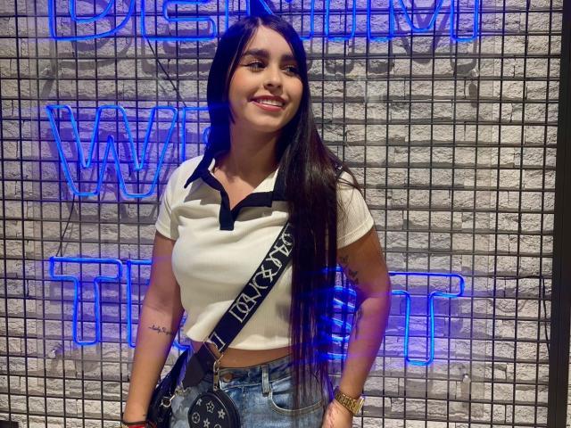 ZoeCastelli with long black hair, white polo, black collar, blue jeans, black bag with "LEVI'S" strap, smiling, tattoo on left arm, brick wall, and blue neon lights.