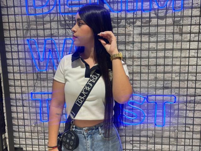 ZoeCastelli with long black hair, white polo, black neck strap, blue jeans, gold watch, hand in hair, black bag, neon blue background, tattoo on left forearm.