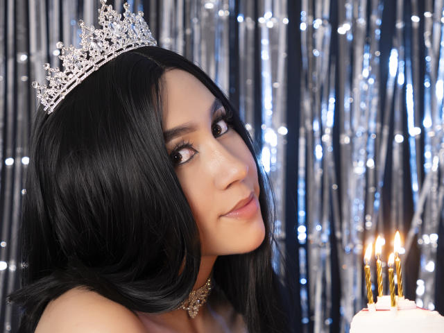 CelesteRusso with long black hair, wearing a silver tiara, looking at the camera with a slight smile, holding a cake with candles in front of her.