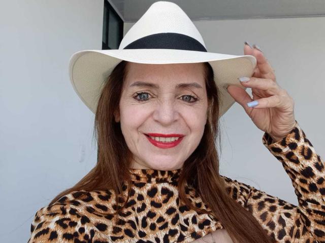 SamantaMature69 con rossetto rosso, che sorride, indossa una maglietta a stampa leopardo e un cappello bianco, tiene il cappello con la mano destra, capelli castani, occhi azzurri, unghie bianche..
