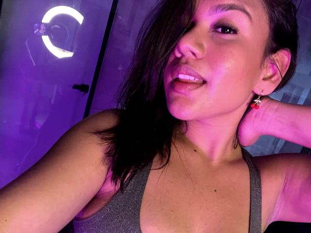AraFerrer with dark hair, pinkish skin, and small earrings, showing off her tits in a gray tank top, tongue out, and hand behind head, under purple light.
