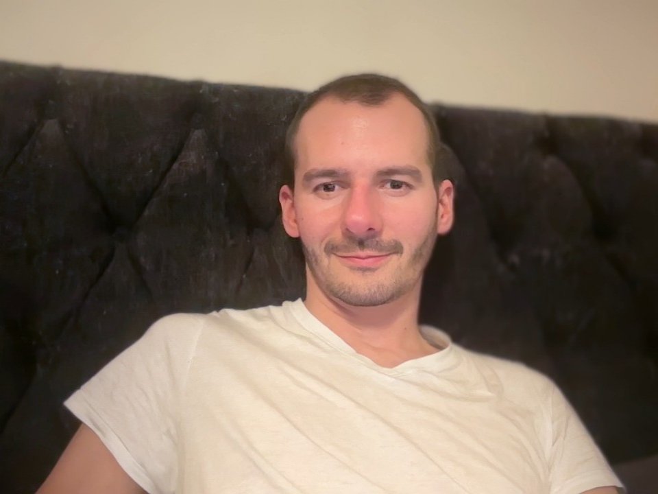 A photograph of ElCapitano with a bald head and light brown hair, short beard, and slight mustache. He's sitting against a black, tufted velvet headboard. ElCapitano is wearing a plain white V-neck t-shirt. His face has a light pinkish tone, and he's smiling slightly. The image is softly lit, with a subtle shadow on the headboard. ElCapitano's skin is fair, and his expression is relaxed.
