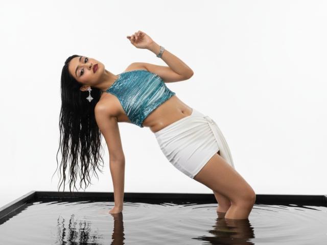 Afrodita, avec ses cheveux noirs et mouillés, portant un haut court vert émeraude et une jupe blanche, est penchée en arrière dans l'eau, montrant ses fesses musclées et sa taille, avec un bracelet à son poignet droit..
