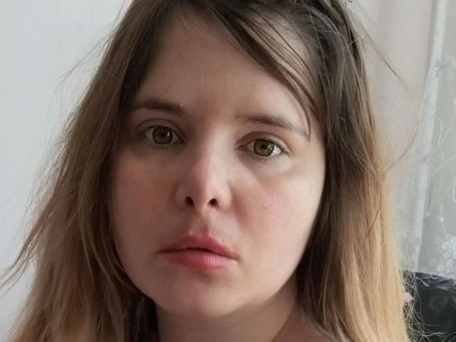 Close-up of ElenaSchatz-hot with light brown, slightly wavy hair falling around her face. She has fair skin, brown eyes, and slightly parted pink lips. Her expression is neutral, with a subtle hint of seriousness. She wears a black hair clip on the left side of her head. The image focuses on her face, with soft light illuminating her features. The background is out of focus, showing a white curtain on the right. The texture of her hair is slightly messy. Her eyes are centered in the frame.