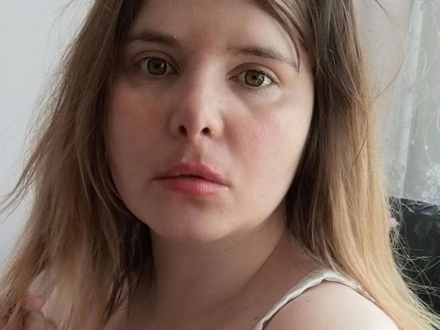 Close-up photograph of ElenaSchatz-hot. She has light skin, brown eyes, and slightly parted pink lips. Her straight, shoulder-length brown hair with blonde highlights frames her face. She wears a white strap top. The image has a soft, natural light, highlighting her facial features. The texture of her skin is smooth with a subtle pink blush. The focus is on her face, which occupies most of the frame. Her expression is neutral, slightly serious.