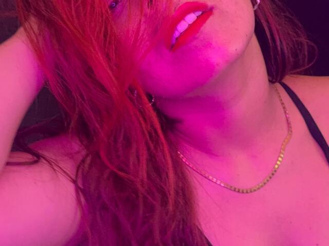 Il viso di MeghanStorm69, con rossetto rosso e capelli rossi bagnati, illuminato da una luce rosa-viola, mostra una collana d'oro e un reggiseno nero, la testa leggermente girata, un sorriso e una mano tra i capelli..