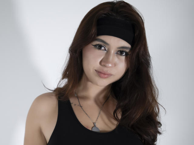 AilenTovar has brown wavy hair, a black headband, black tank top, and a necklace with a pendant, showing off her tits and a subtle, sexy smile.