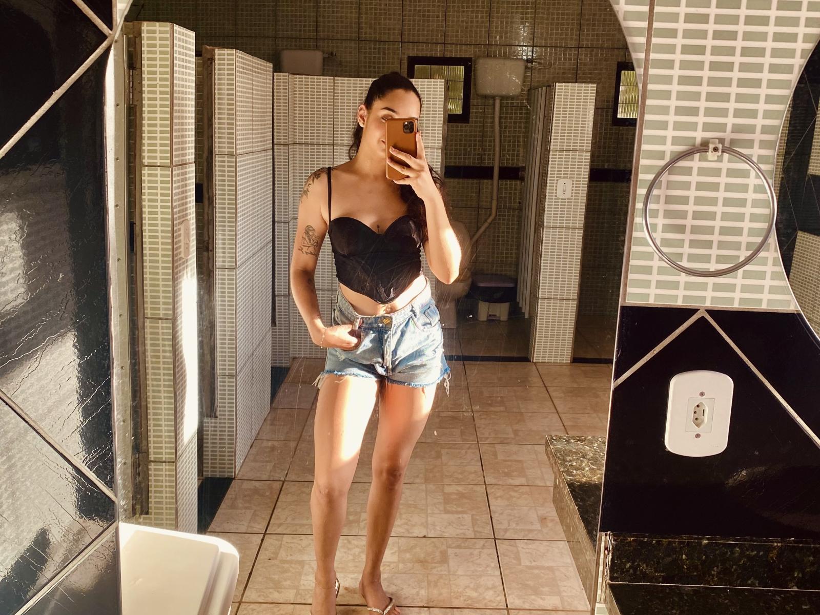 DrySecret con tatuaggi, indossa una maglietta nera con lacci che lascia intravedere il seno e pantaloncini di jeans, in piedi in un bagno, che si fa un selfie allo specchio con il suo telefono, in piena luce solare..