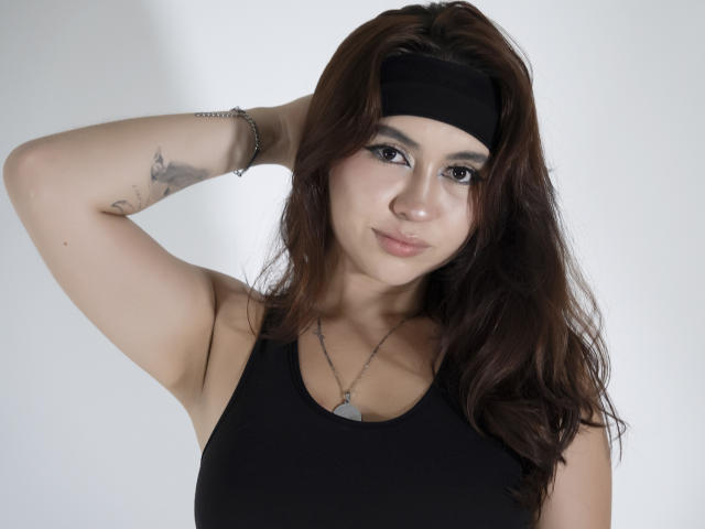 AilenTovar with wavy brown hair, black headband, black tank top, and arm tattoo, raising arm, showing small tits, subtle makeup, necklace, and silver bracelet.