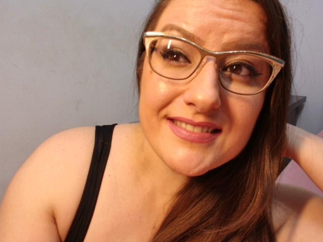 BadAngel with glasses, smiling, brown hair, pink lips, black tank top, visible tits, fair skin, hand behind head, slightly wavy eyebrows, light blue background.