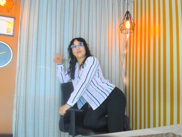 JenniZeller with curly black hair, blue glasses, striped white shirt, black pants, leaning on chair, smiling, small breasts, medium build, in a brightly lit room.