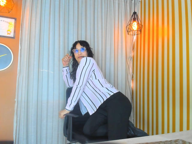 JenniZeller with curly black hair, blue mirrored sunglasses, striped white shirt, black pants, kneeling on chair, looking back, orange wall, hanging light, white curtain.