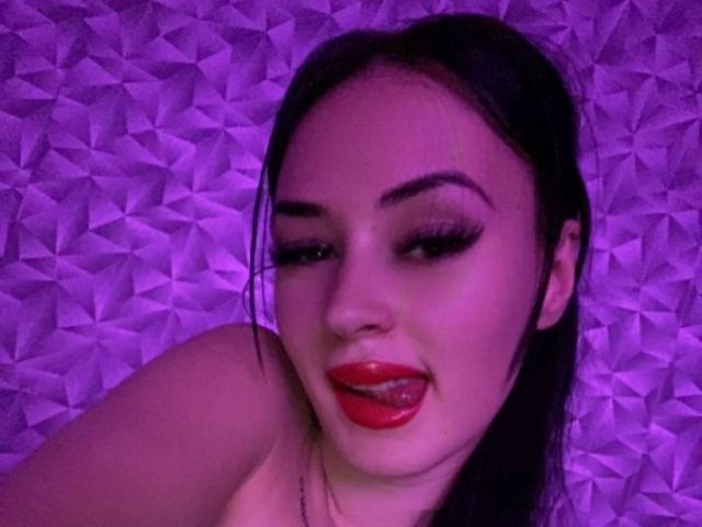 Ruby Sinclair, com cabelo escuro, batom vermelho e a língua de fora, tem sobrancelhas marcantes e cílios longos, contrastando com padrões geométricos roxos. Seus ombros nus são visíveis..