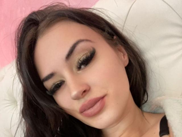 Ruby Sinclair tem cabelo castanho escuro, olhos verdes com cílios pretos grossos e lábios rosados cheios. Ela está olhando para baixo com um leve sorriso, deitada em um travesseiro branco..