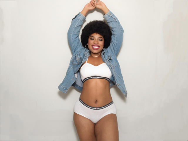 NatashaSparks with dark skin, big afro, smiling, wearing a denim jacket, white bra, and white panties with black trim, arms raised, standing, showing her tits and ass.