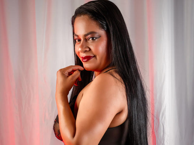 TeresaHearts with long black hair, red lipstick, and black lingerie, smiling at the camera, holding her chin with a hand, her tits partially visible, red lighting.