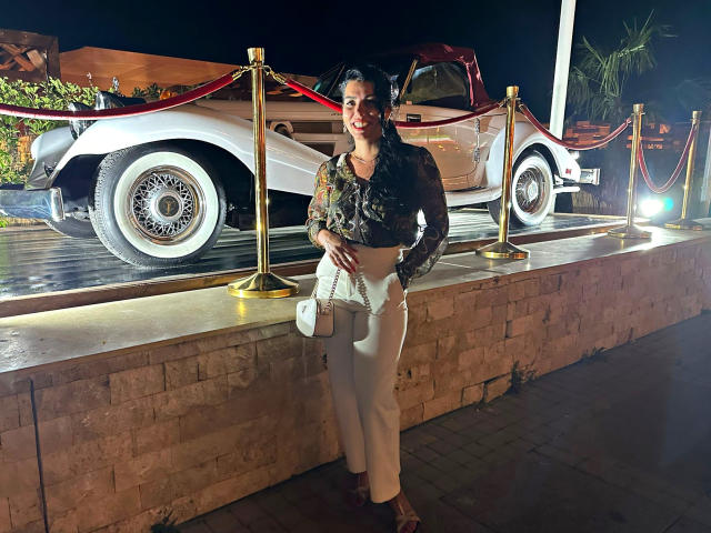 TonyaAntonyaLips stands in front of a white car, wearing a black floral top, white pants, and a white purse, smiling with short dark hair, and a medium ass.