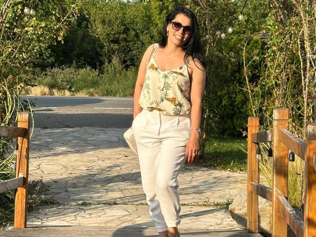 TonyaAntonyaLips with long black hair, wearing sunglasses, a floral tank top, and white pants, smiling on a stone pathway, holding a white bag, surrounded by greenery.