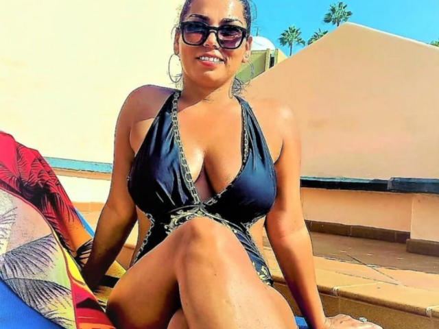 TonyaAntonyaLips with big tits in a black halter dress, sitting on a roof, sunglasses on, smiling, knees together, sunlit, blue sky, palm trees, colorful pillow in background.