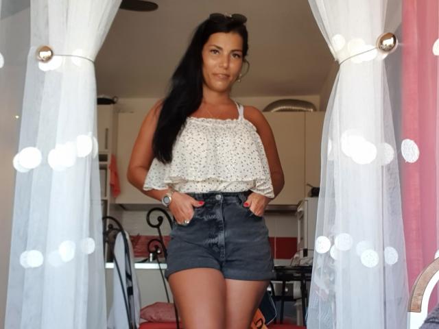 TonyaAntonyaLips with long black hair, wearing a white speckled sleeveless top and dark denim shorts, standing in front of white curtains with circular lights. She's smiling.