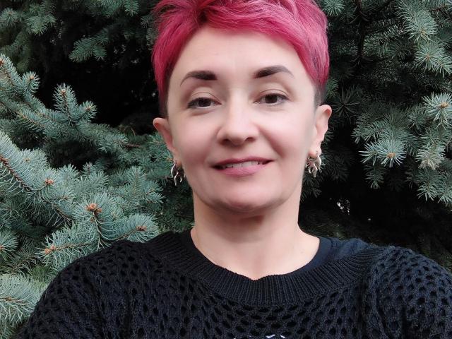 ChristinaRainforce with pink hair, black patterned sweater, and earrings, smiling, standing in front of a green pine tree. Her tits are small, and she's cute.