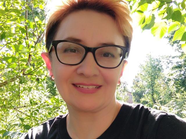 ChristinaRainforce with short orange hair, black glasses, and a black shirt, smiling with a small tooth gap, green leafy background, bright sunlight.